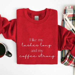 May include: Red sweatshirt with white text that reads "I like my lashes long and my coffee strong". The sweatshirt is folded and laid on a white surface with a pair of red and white checked pyjama trousers.