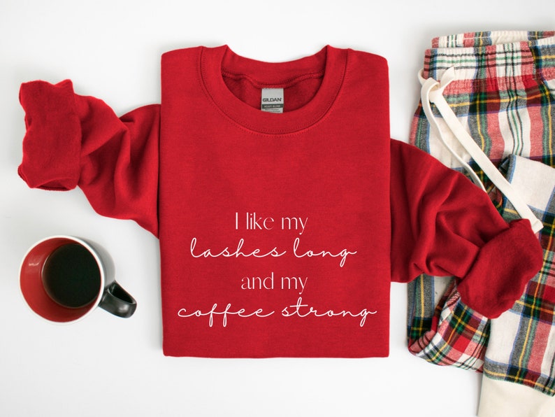 May include: Red sweatshirt with white text that reads "I like my lashes long and my coffee strong". The sweatshirt is folded and laid on a white surface with a pair of red and white checked pyjama trousers.