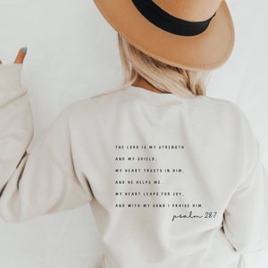 Puede incluir: Una sudadera de color crema con una cita en negro del Salmo 28:7. La cita dice: "El Se&ntilde;or es mi fortaleza y mi escudo. Mi coraz&oacute;n conf&iacute;a en &eacute;l, y &eacute;l me ayuda. Mi coraz&oacute;n se regocija, y con mi canto lo alabo. Salmo 28:7"