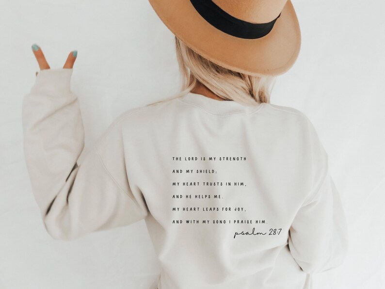 Puede incluir: Una sudadera de color crema con una cita en negro del Salmo 28:7. La cita dice: "El Se&ntilde;or es mi fortaleza y mi escudo. Mi coraz&oacute;n conf&iacute;a en &eacute;l, y &eacute;l me ayuda. Mi coraz&oacute;n se regocija, y con mi canto lo alabo. Salmo 28:7"