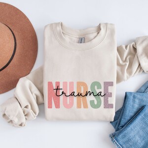 May include: A beige sweatshirt with the words "nurse trauma" in colorful, block letters. The word "nurse" is in pink, orange, green, and purple. The word "trauma" is in a handwritten font. A brown hat and blue jeans are in the background.