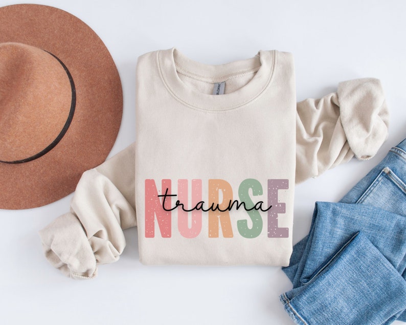 May include: A beige sweatshirt with the words "nurse trauma" in colorful, block letters. The word "nurse" is in pink, orange, green, and purple. The word "trauma" is in a handwritten font. A brown hat and blue jeans are in the background.