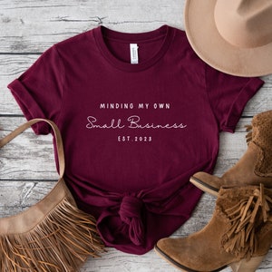 May include: Burgundy t-shirt with the text "Minding My Own Small Business Est. 2023" in white. The shirt is laid out on a wooden surface with a brown hat, a brown fringed bag, and brown boots.