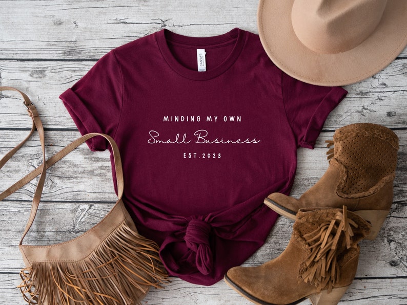 May include: Burgundy t-shirt with the text "Minding My Own Small Business Est. 2023" in white. The shirt is laid out on a wooden surface with a brown hat, a brown fringed bag, and brown boots.