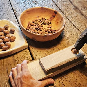 Könnte beinhalten: Ein helles Nussknacker-Werkzeug aus Holz mit einer kreisförmigen Öffnung, ein Metallhammer, eine herzförmige Holzschale gefüllt mit ganzen Walnüssen und eine runde Holzschale mit Walnussschalen, angeordnet auf einer rustikalen Holzoberfläche.
