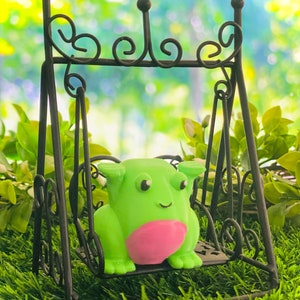 May include: A green frog with a pink belly sits on a black metal swing set. The swing set is in a grassy area with a blurred green background.