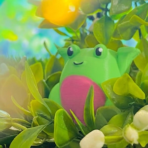 May include: A green frog figurine with pink accents sits in a bed of green leaves and white flowers. The frog has large, black eyes and a small, white smile.