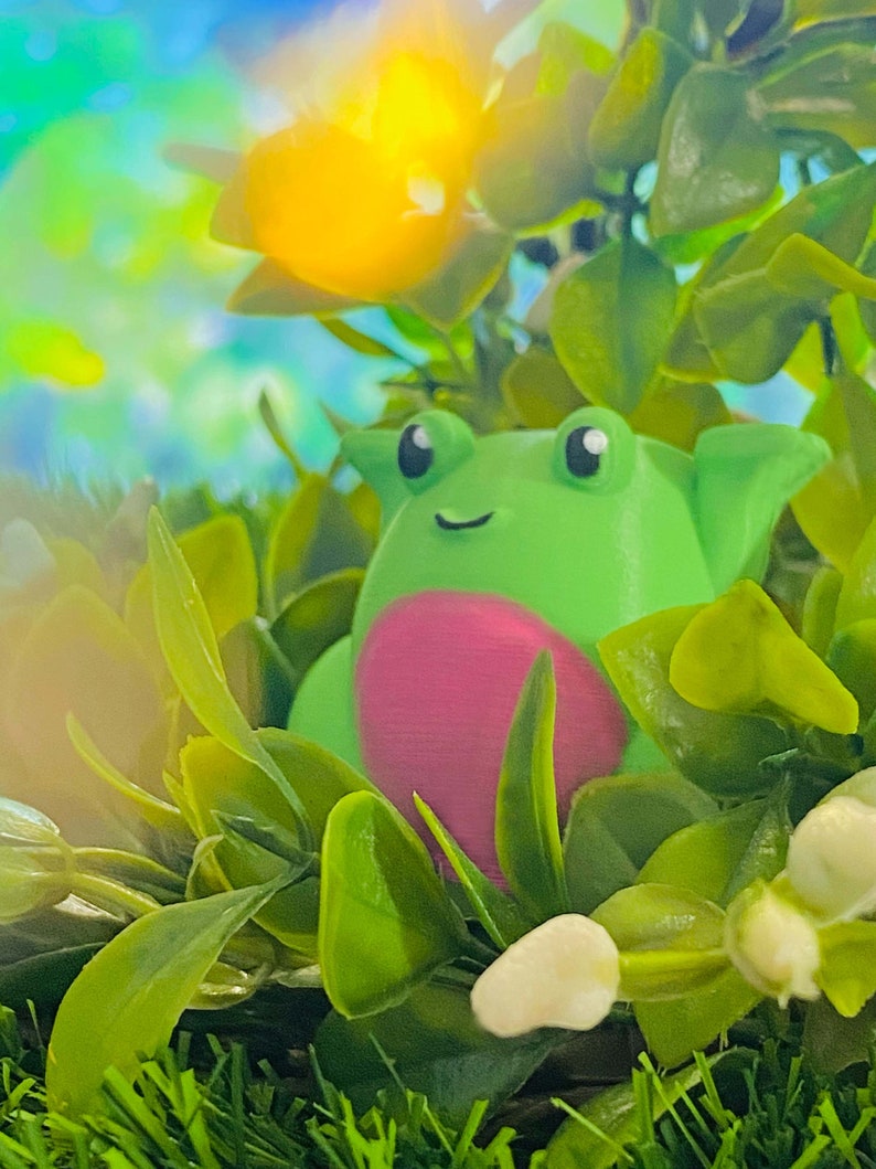 May include: A green frog figurine with pink accents sits in a bed of green leaves and white flowers. The frog has large, black eyes and a small, white smile.