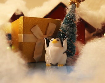 May include: A white penguin figurine with yellow beak and black eyes stands in front of a gold gift box with a white ribbon and a small green Christmas tree.