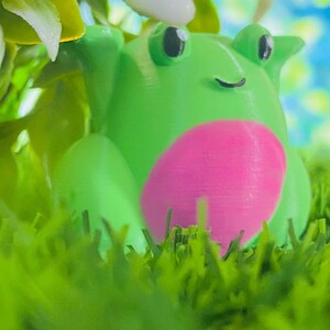 May include: A green frog figurine with a pink belly, sitting in green grass. The frog has black eyes and a small smile.