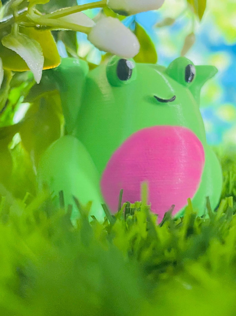 May include: A green frog figurine with a pink belly, sitting in green grass. The frog has black eyes and a small smile.