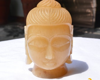 May include: A carved light orange stone sculpture of a Buddha head featuring a smooth, polished finish, closed eyes, and stylized hair patterns. The sculpture displays subtle natural color variations and is positioned upright on a flat surface.
