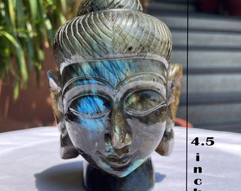 May include: A hand-carved labradorite stone sculpture of a head with a serene expression and detailed hair texture. The stone displays iridescent blue flashes across the surface. It measures 4.5 inches in height and 3.7 inches in width.