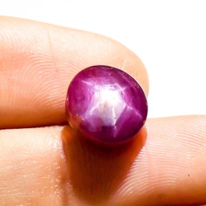 May include: A polished, oval-shaped ruby gemstone. The gem is a deep magenta color with a six-rayed star effect visible on its surface. The star effect is created by light reflecting off of inclusions within the stone.