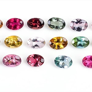 Puede incluir: Una variedad de gemas de corte ovalado en varios colores, incluyendo tonos de marrón, rosa, verde y azul. Las gemas están dispuestas sobre una superficie blanca, mostrando sus facetas pulidas y tonos vibrantes. Ideal para la creación de joyas.