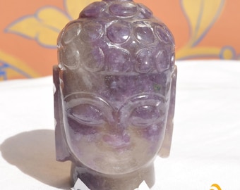 May include: A carved purple amethyst stone sculpture in the shape of a Buddha head. The figure features stylized hair curls, closed eyes, and elongated earlobes, with a polished, translucent surface showing natural mineral inclusions throughout.
