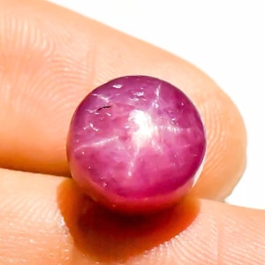 May include: A round, cabochon-cut star ruby gemstone with a deep magenta-red color. The surface displays a distinct six-rayed star effect, known as asterism, which appears as a bright white light reflection centered on the polished, smooth dome.