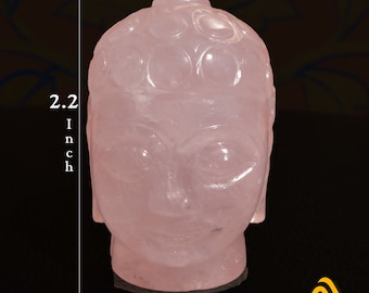 May include: A light pink rose quartz carving of a Buddha head with stylized hair curls. The sculpture features a smooth, polished surface and measures 2.2 inches in height and 1.6 inches in width as indicated by the white measurement lines.