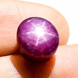 Puede incluir: Una gema pulida, de forma ovalada, de color magenta intenso. La gema presenta un patrón de estrella de seis rayos en su superficie, creando un efecto de estrella. La piedra se sostiene en una mano, mostrando su color y brillo.