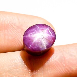 May include: A polished, oval star ruby gemstone. The gem is a deep purplish-red colour with a six-rayed star pattern visible on its surface. The star effect is caused by light reflecting off inclusions within the stone.