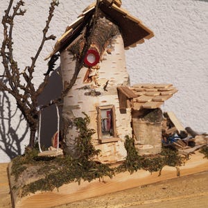 Puede incluir: Una casa rústica en miniatura hecha de corteza de abedul, madera y musgo. La casa presenta un tejado de tejas de madera, una pequeña ventana y un acento circular rojo. Un pequeño árbol y un columpio están unidos. La casa está sobre una base de madera.