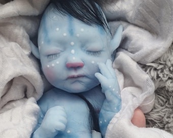 May include: A blue silicone baby doll with pointed ears, dark hair, and white spots on its skin. The doll is wrapped in a white fabric and features the text Dux & Binky SILICONE BABIES by Louise Jones & Beverley Kimble printed on its torso.