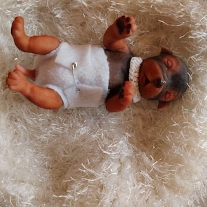 Op de afbeelding: Een levensechte miniatuur puppy pop, liggend op zijn rug, met een witte luier vastgezet met een veiligheidsspeld en een gehaakte kraag. De puppy heeft bruine en zwarte vacht, gesloten ogen en ligt op een zacht, wit oppervlak.