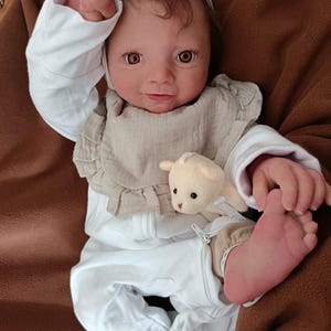 Può includere: Una bambola reborn realistica con occhi marroni e capelli scuri, che indossa una tutina bianca a maniche lunghe, una bavaglina beige con volant e una fascia beige annodata abbinata. La bambola tiene in mano un piccolo orsetto di peluche crema.