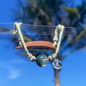 Op de afbeelding: Een groene stenen armband met een bruin leren bandje en een zilveren bedel. De armband hangt aan een doorzichtige plastic staaf tegen een blauwe lucht met een palmboom op de achtergrond.