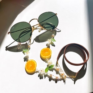 May include: A gold-tone glasses chain with a brown leather cord and white pearl accents. The chain features small, realistic-looking orange slices and white flower charms. The chain is attached to a pair of round, green-tinted sunglasses.