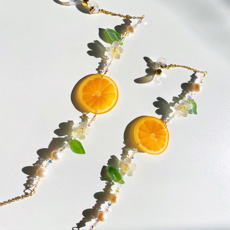 May include: A pair of gold-tone glasses chains with orange citrus fruit charms and white flower accents. The chains are adorned with clear and iridescent beads.