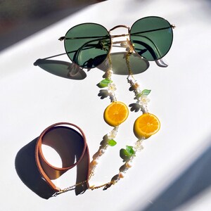 May include: A gold chain with white pearls and green leaves, accented with orange lemon slices, connects to brown leather straps. The chain is designed to hold glasses.