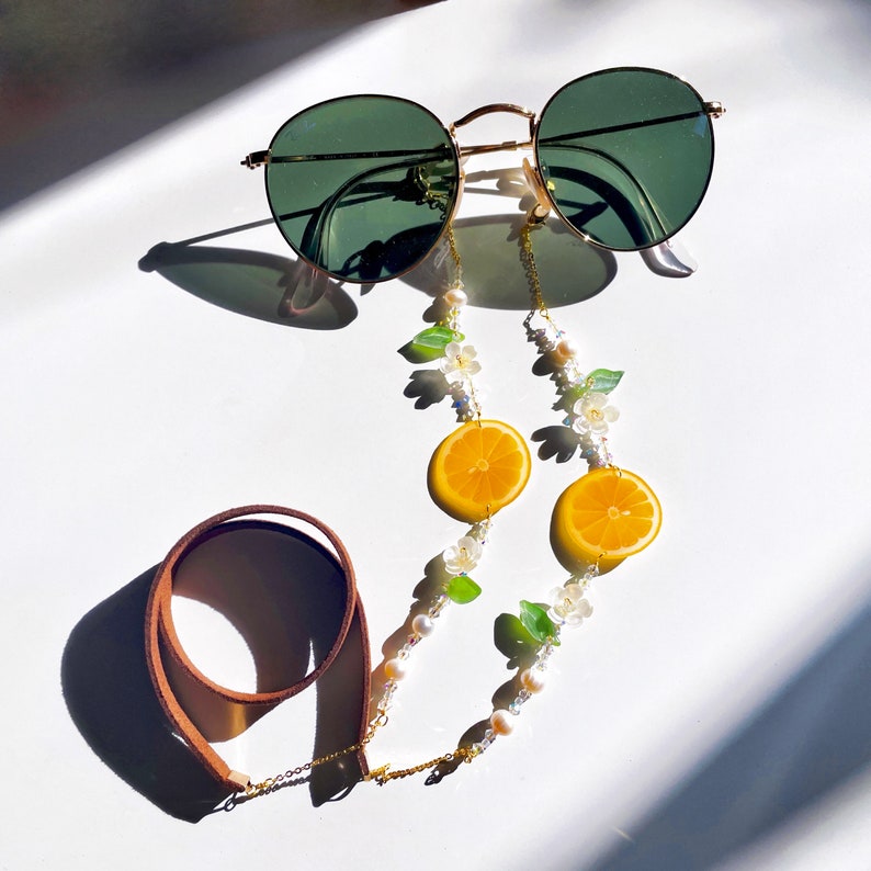 May include: A gold chain with white pearls and green leaves, accented with orange lemon slices, connects to brown leather straps. The chain is designed to hold glasses.