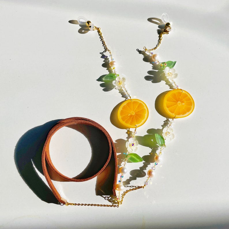 May include: A gold chain with lemon slices, white flowers, and green leaves. The chain has two gold clasps and a brown leather strap.