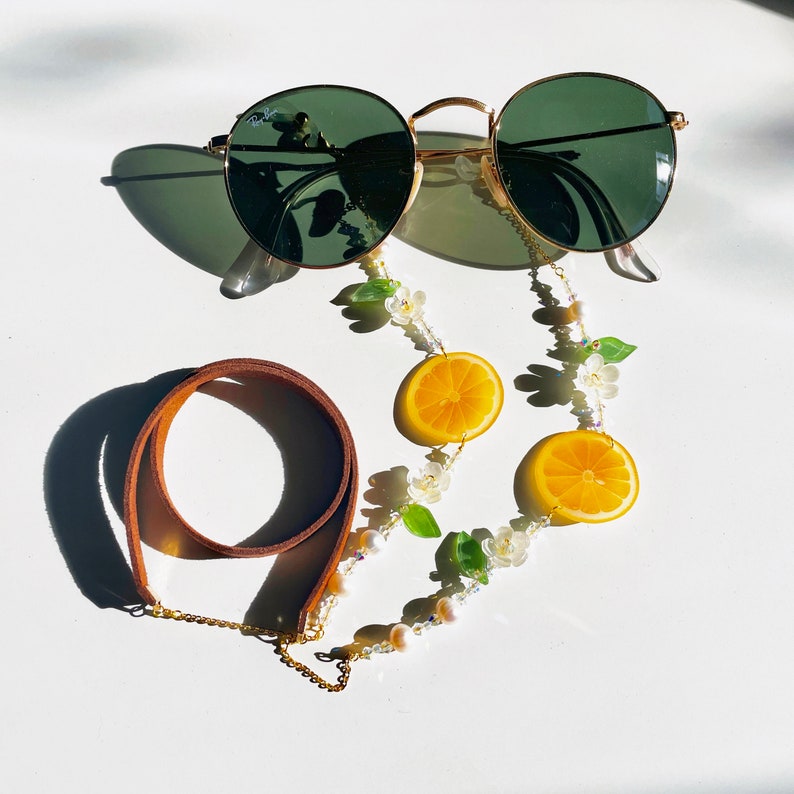 May include: A brown leather strap with a gold chain and white pearl beads. The chain is attached to a pair of green and gold glasses with a lemon slice charm.