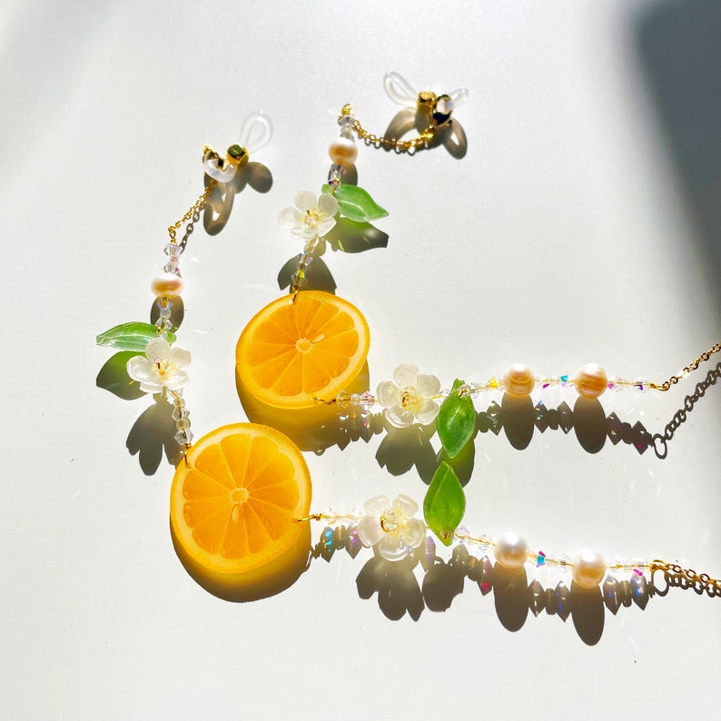 May include: A gold chain with white and green flower charms, two lemon slices, and pearl beads. The chain has two gold clasps.