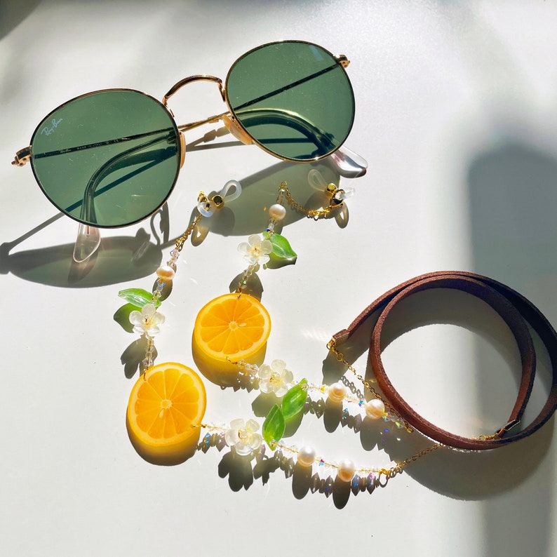 May include: A pair of green sunglasses with a gold frame and a chain with lemon slices and white flowers attached. The chain is connected to a brown leather strap.