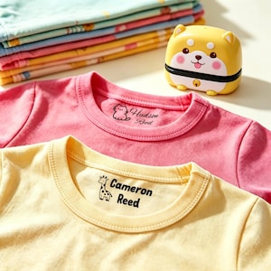 以下が含まれることがあります： 猫のアイコンとHudson Reedの文字が入ったピンクのTシャツ、キリンのアイコンとCameron Reedの文字が入った黄色のTシャツ、折りたたまれたカラフルなTシャツの山、そして犬の顔がデザインされた黄色の動物型プラスチックスタンプ。