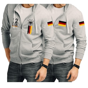 Könnte beinhalten: Zwei graue Kapuzenpullover mit Reißverschluss und schwarz-gelben deutschen Flaggen-Designs. Ein Hoodie hat die Nummer 12 und das Wort "GER" auf der Vorderseite. Der andere Hoodie hat die deutsche Flagge auf dem linken Ärmel und dem rechten Ärmel.