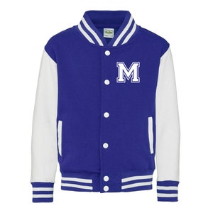 Op de afbeelding: Een koninklijk blauwe en witte varsity jas met een witte letter "M" op de borst. De jas heeft een knoopsluiting en witte geribbelde manchetten en tailleband.