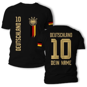 Può includere: Una t-shirt nera a maniche corte con grafiche dorate. Il davanti mostra la scritta DEUTSCHLAND, il numero 10 e un emblema dell'aquila tedesca con quattro stelle. Il retro presenta DEUTSCHLAND, il numero 10 e la scritta DEIN NAME in oro.