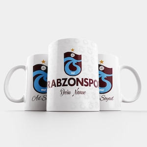 Può includere: Tre tazze in ceramica bianca con un logo blu e marrone che presenta un'onda e una stella. Il logo dice "Trabzonspor" e "1967". Le tazze sono personalizzate con il testo "Dein Name", "Ad Soyad" e "Soyad".