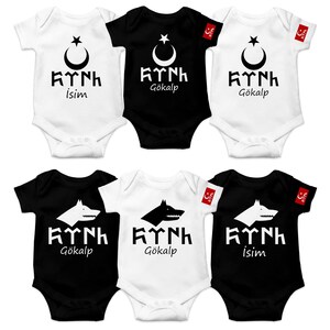 Op de afbeelding: Zes baby rompertjes in zwart en wit met een halve maan en ster ontwerp, de woorden "KINH" en ofwel "Gökalp" of "İsim" gedrukt op elk rompertje.