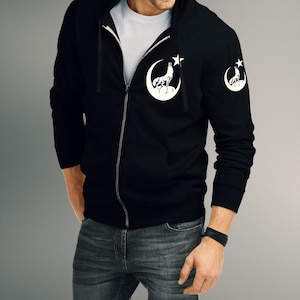 Op de afbeelding: Een zwarte hoodie met rits met een wit wolvenontwerp dat huilt naar een ster en een halve maan op de voorkant en een kleinere versie van het ontwerp op de linkermouw.