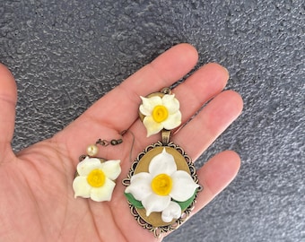 Può includere: Un set di gioielli fatti a mano con un ciondolo e orecchini, ognuno ornato da un motivo di narciso bianco e giallo. Il ciondolo è di forma ovale con una cornice in bronzo ornata. Gli orecchini hanno un accento di perla.