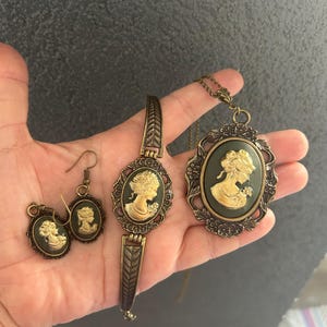 Può includere: Un set di gioielli vintage con design a cammeo. Il set include orecchini, un braccialetto e una collana con pendente. Ogni pezzo ha uno sfondo ovale verde scuro con un profilo bianco di donna. Le montature e il braccialetto sono color bronzo.