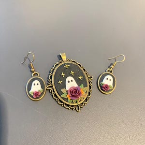 Op de afbeelding: Een sieradenset met Halloween-thema, bestaande uit een hanger en oorbellen. De goudkleurige hanger en oorbellen hebben elk een wit spookontwerp met rozen en sterren op een zwarte achtergrond. Een charmant en griezelig accessoire.