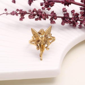 May include: A gold-colored ring featuring a fairy design with outstretched wings. The fairy is depicted in a standing pose, with intricate details in the wings and body. The ring is set against a white surface with a sprig of dried purple flowers.