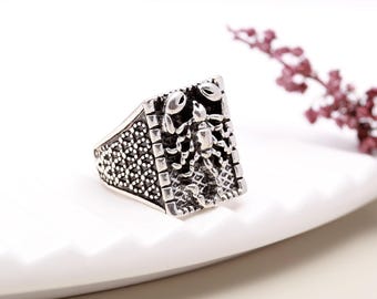 Puede incluir: Anillo plateado con una cara rectangular que presenta un diseño detallado de escorpión. La banda del anillo tiene un patrón texturizado, y el escorpión está intrincadamente detallado. El anillo se muestra sobre una superficie blanca.