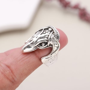 May include: A silver-colored ring with a detailed eagle head design. The ring is worn on a finger and features intricate carvings, including the eagle's eye and feathers. The ring is likely made of metal.